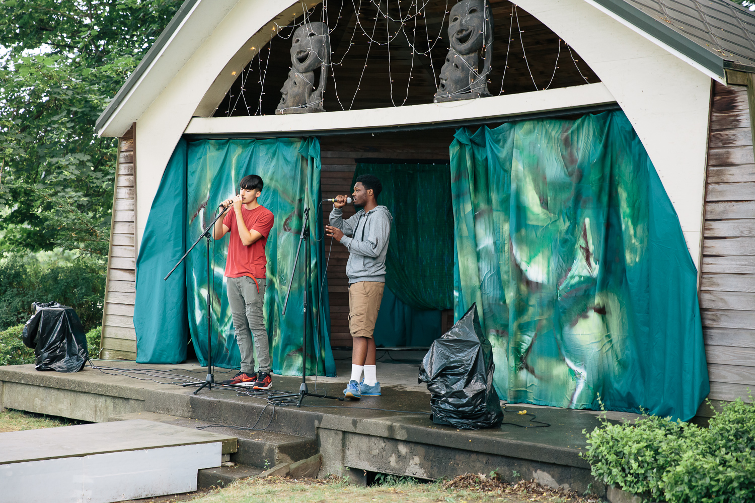 Two musicians stand on a wood stage playing instruments in front of green backdrop.