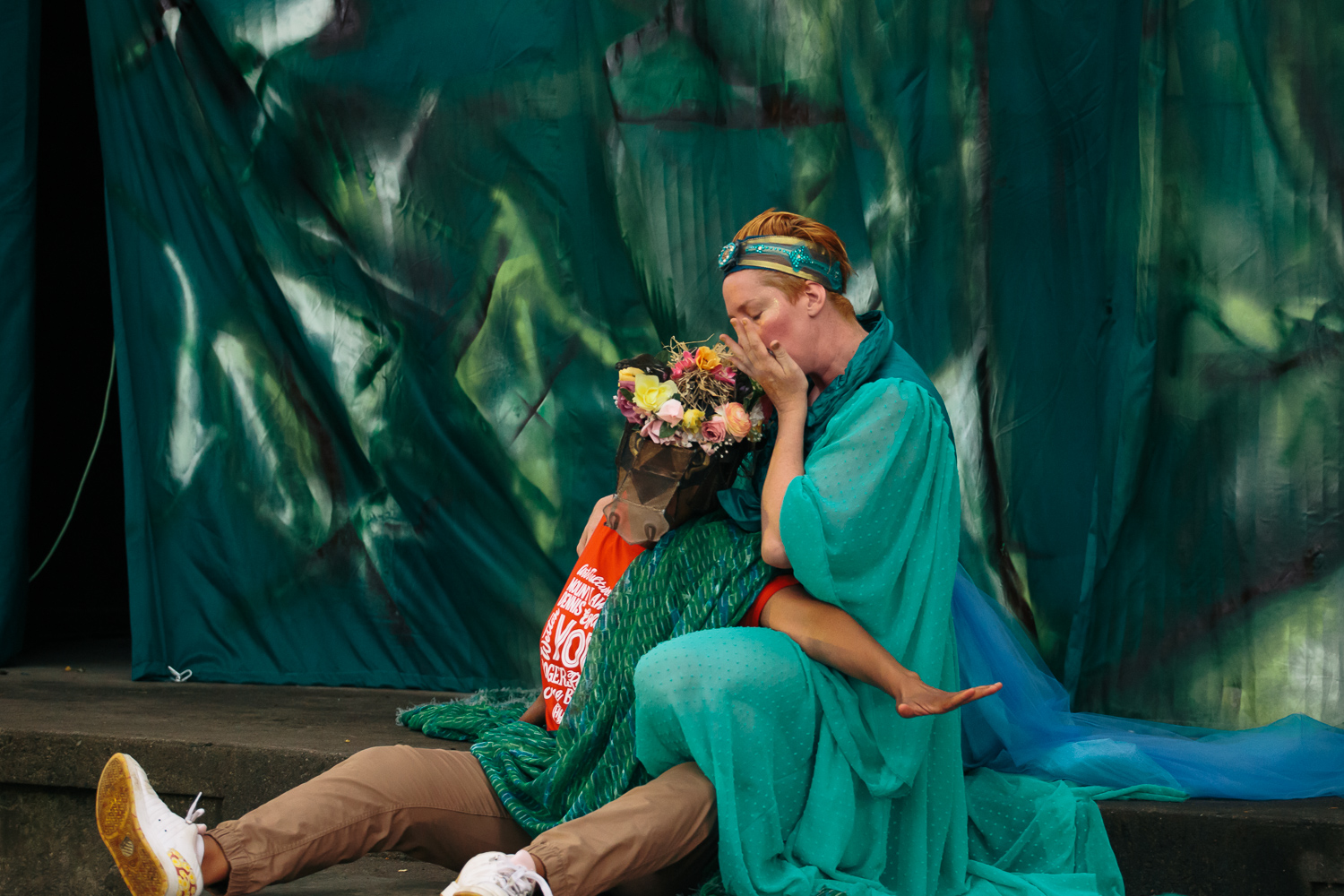 A woman in a green costume sits on a stage with a child who is wearing a crown of flowers.