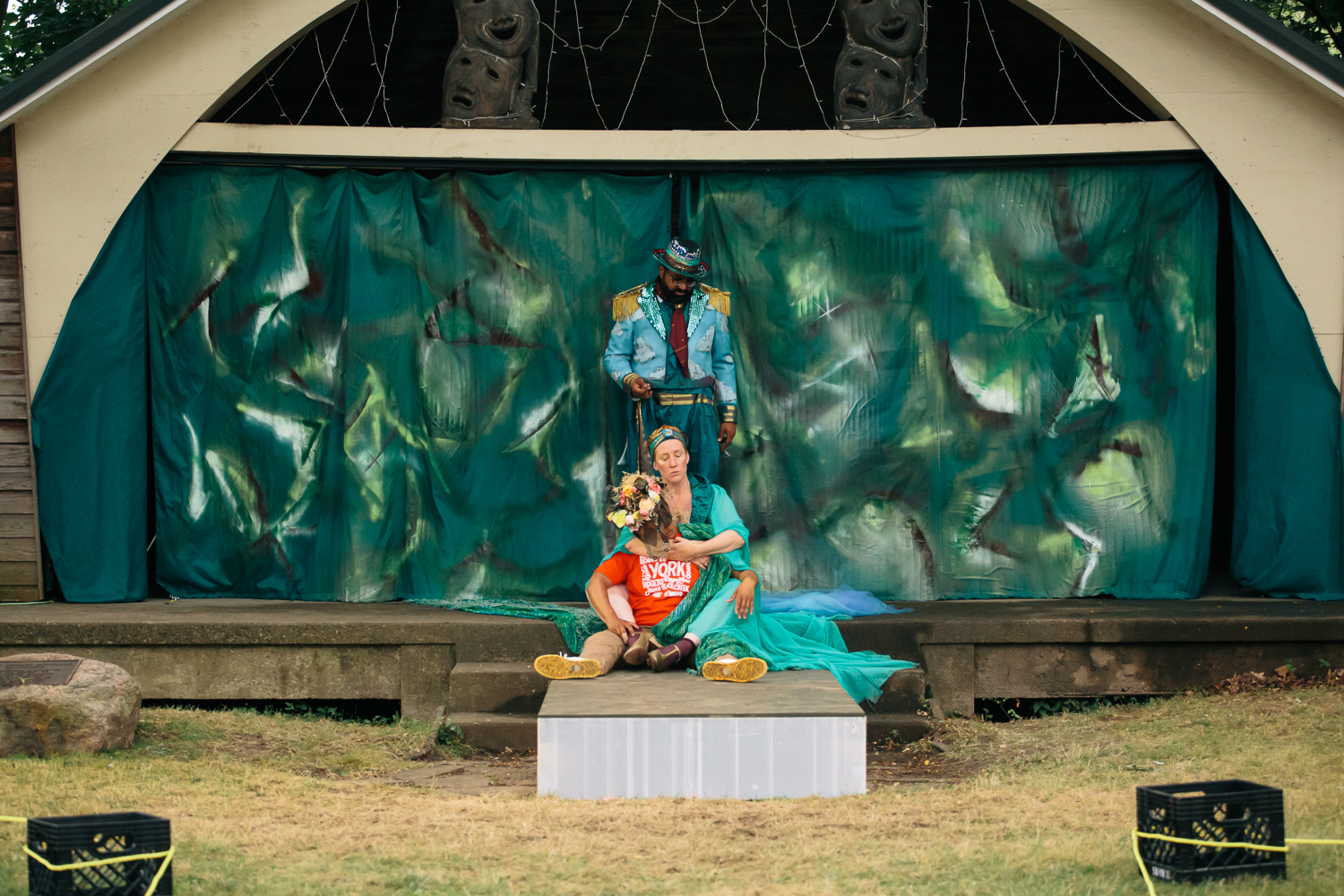 Three performers in costumes stand in the middle of a raised wooden stage with a green backdrop