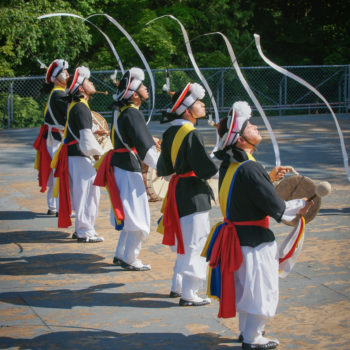 Five Men In Colourful Costumes Play The Drums As They Dance And Move Long Ribbons Attached To Their Hats.