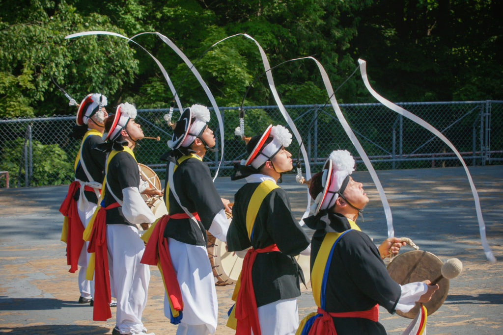 Five men in colourful costumes play the drums as they dance and move long ribbons attached to their hats.