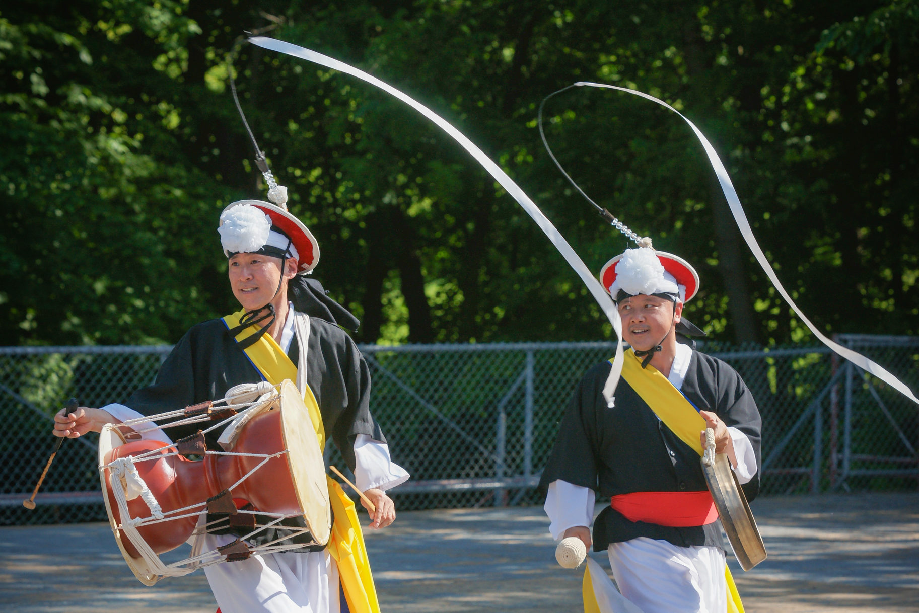 Two men in colourful costumes play the drums as they dance and move long ribbons attached to their hats.