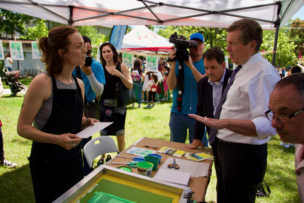 Mayor John Tory stands in front of an artist and learns how to create a silkscreen print on a table.