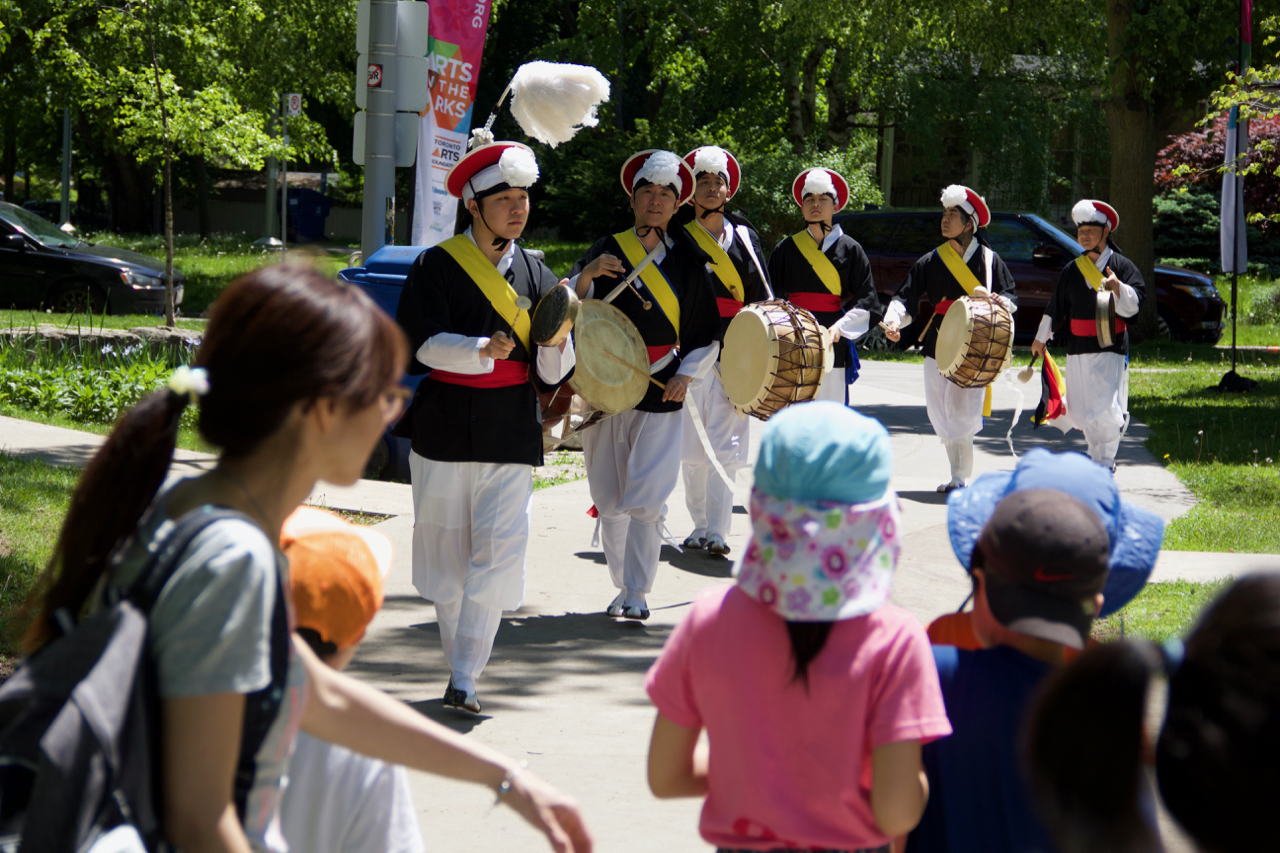 A parade of men playing drums in colourful costume walk down a sidewalk.