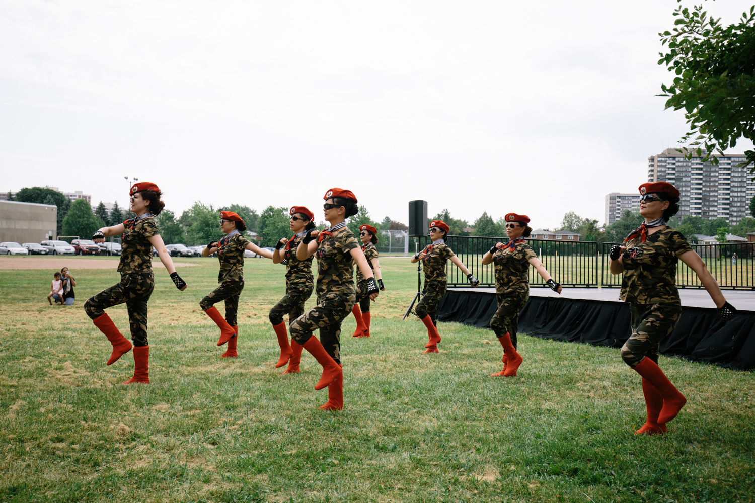 A group of seven dancers in dark costumes and red boots dance on the grass in front of the stage.