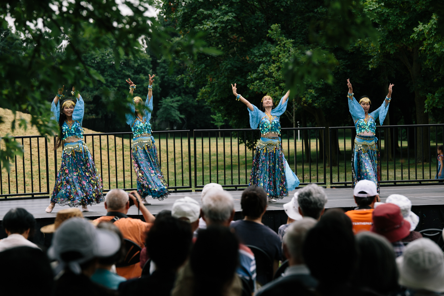 Four women in blue costumes dance on a raised stage while audience members watch.