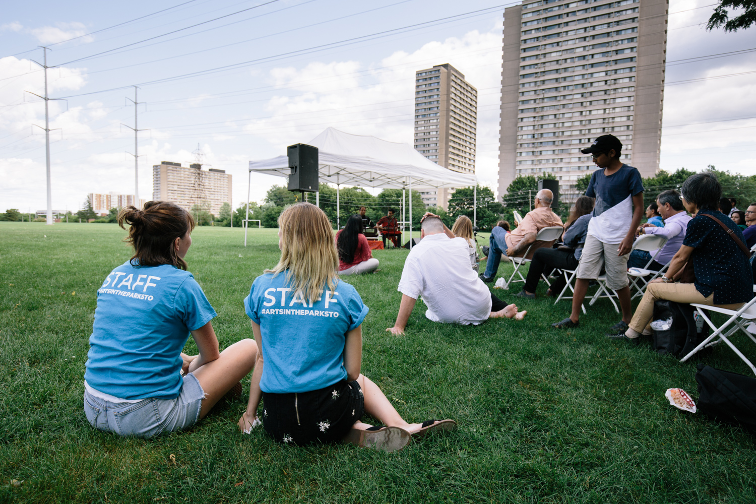 Audience members sit on the grass and in chairs. Two Arts in the Parks staff sit in the foreground wearing blue shirts.
