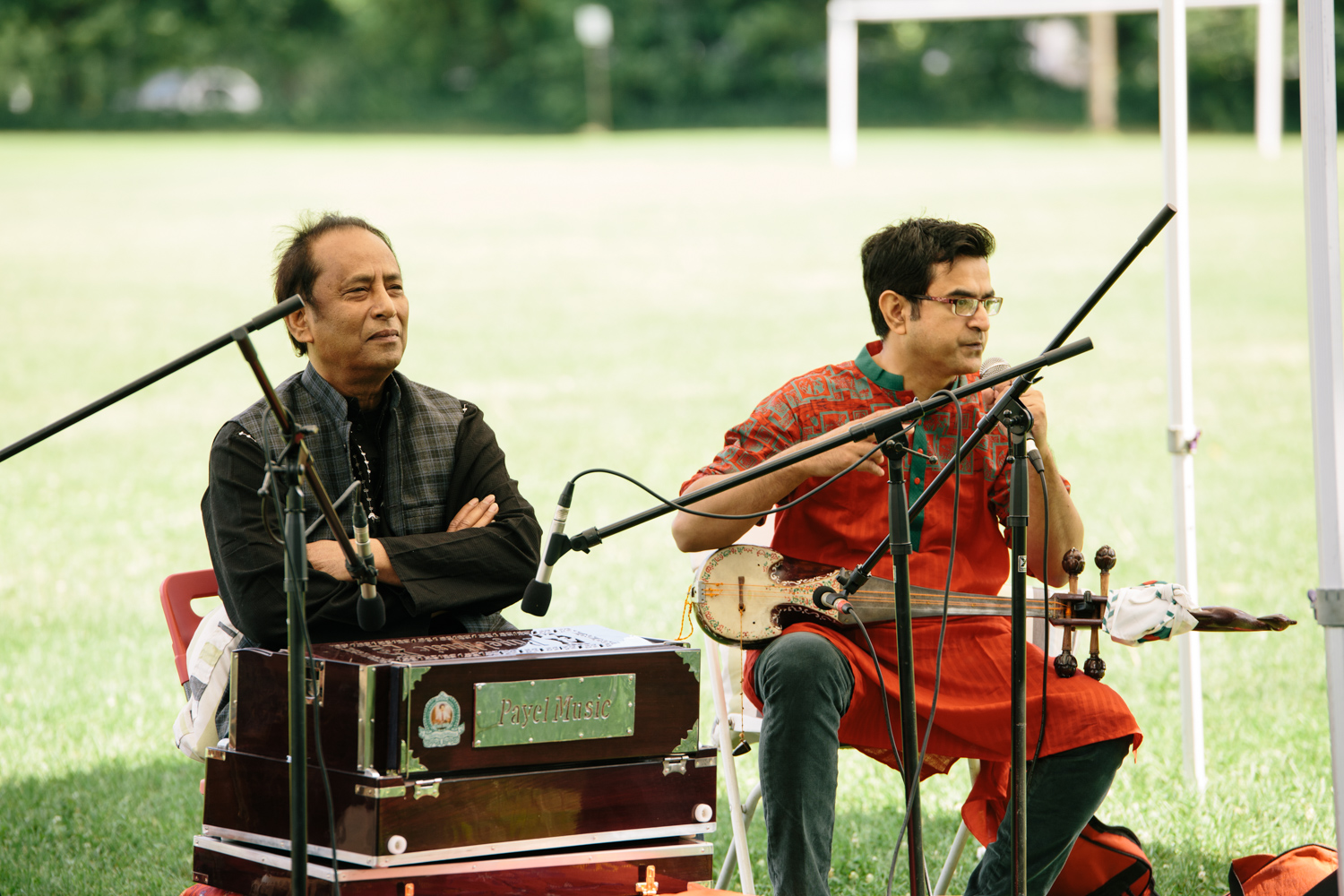 Two men perform different instruments under a tent in a field.