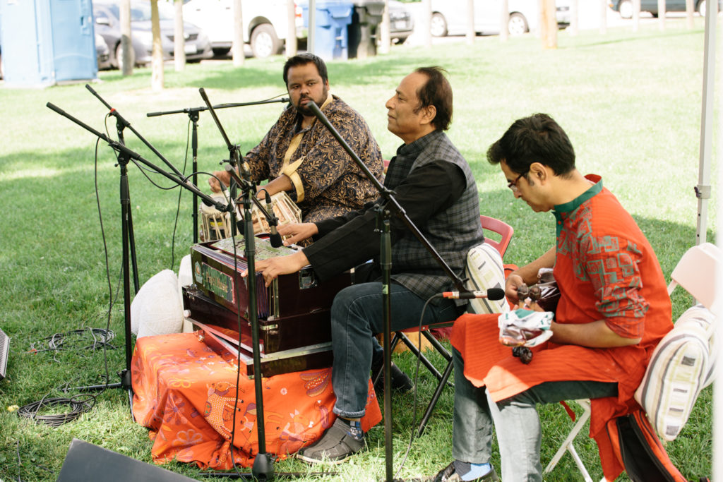 Three men sing and play instruments as they are seated under a tent.