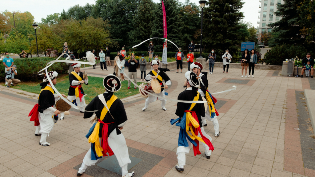 Five drummers dance in a circle. They play handheld instruments while long white ribbons from their hats move in the wind.