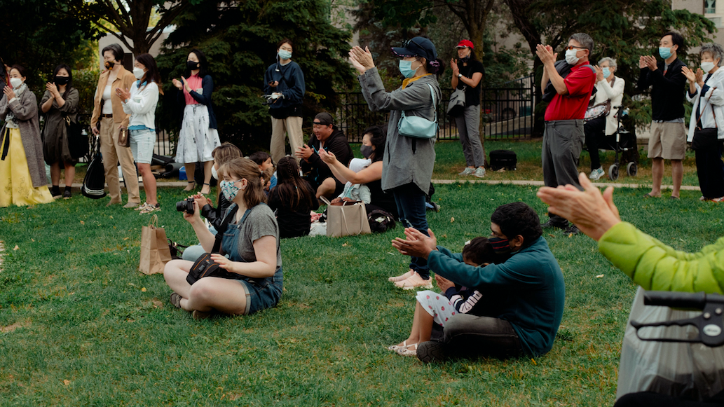 Audience members sit and stand on the grass and clap after a performance.
