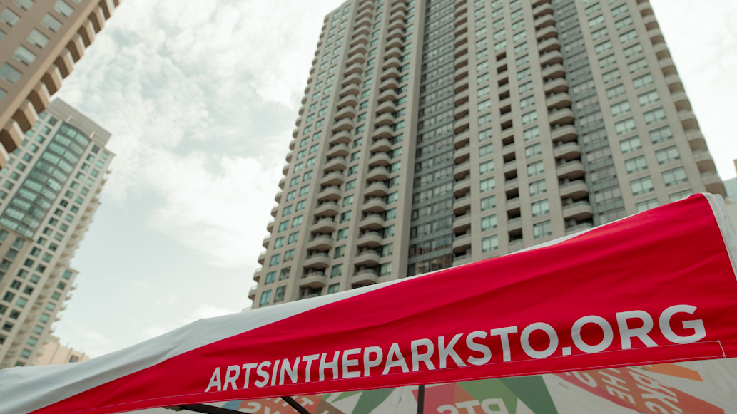 A large condominium tower stands in the background. In front is the side of a tent which is pink and reads "artsintheparksto.org".