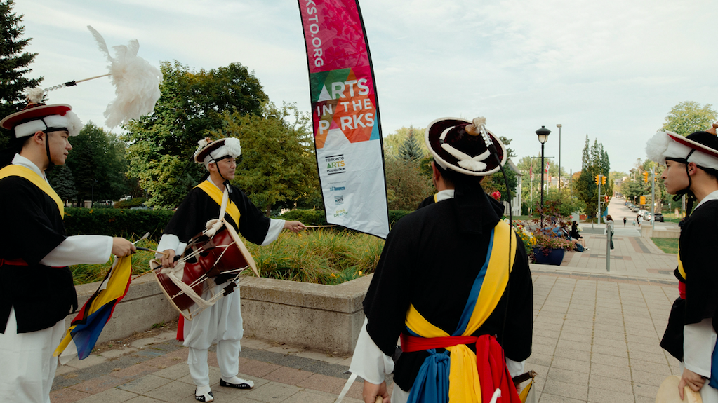 Four drummers in costume stand in a circle with a pink flag in the background.