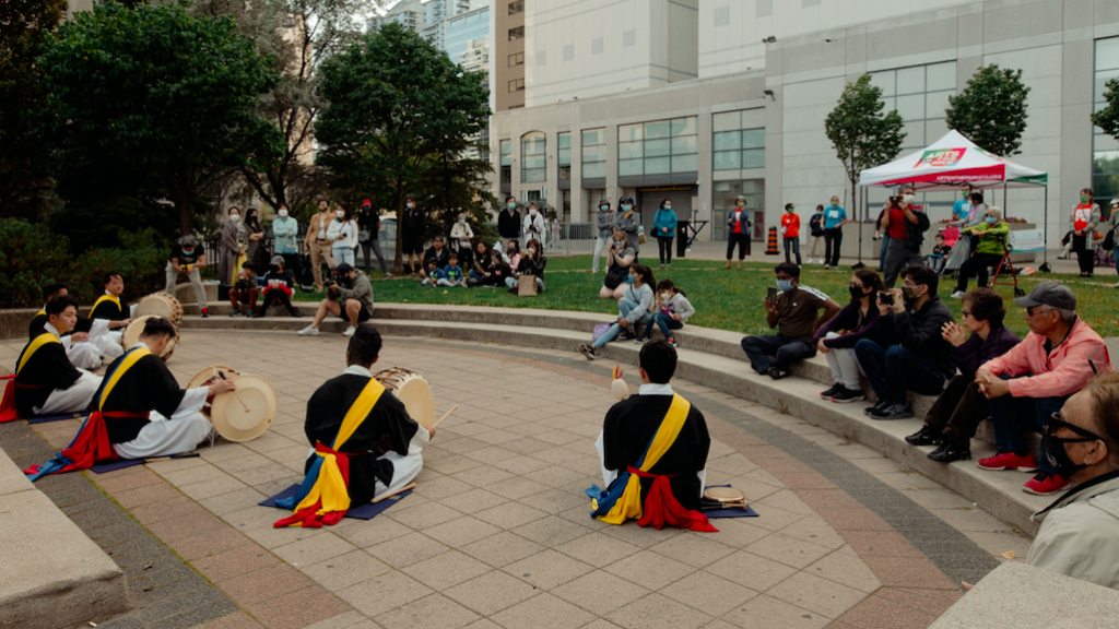 Five drummers in costume sit in a semi circle on the pavement. They each play a handheld percussion instrument while audience members watch from the grass.