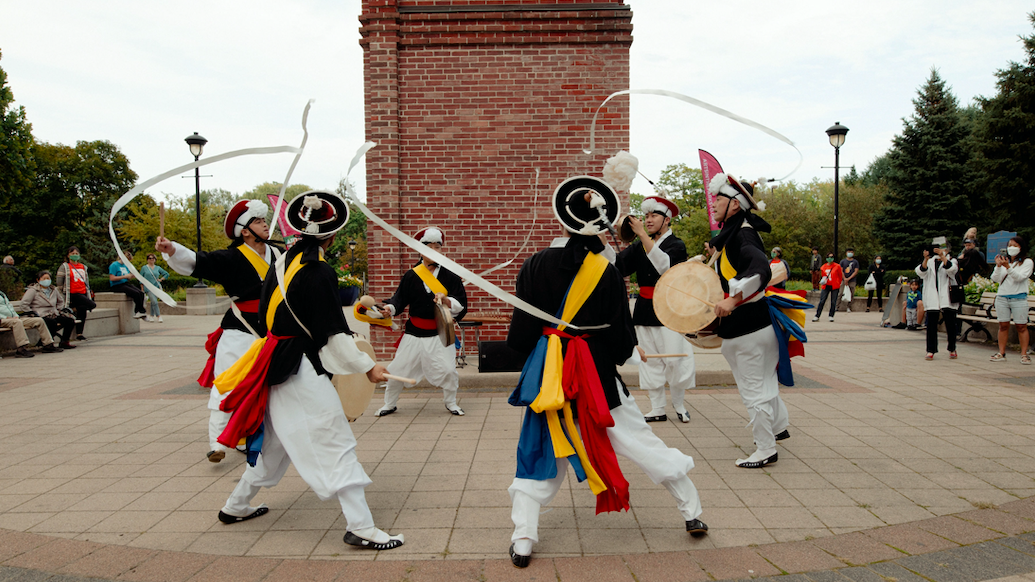 Five drummers dance in a circle. They play handheld instruments while long white ribbons from their hats move in the wind.