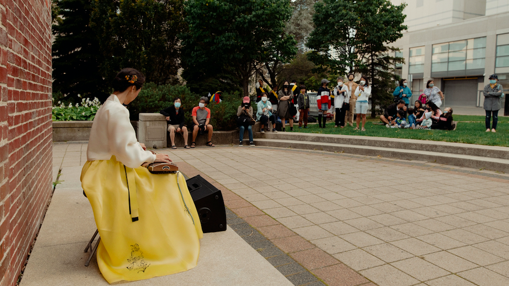 A woman sits in a yellow dress playing a stringed instrument. Audience members watch her from the grass.