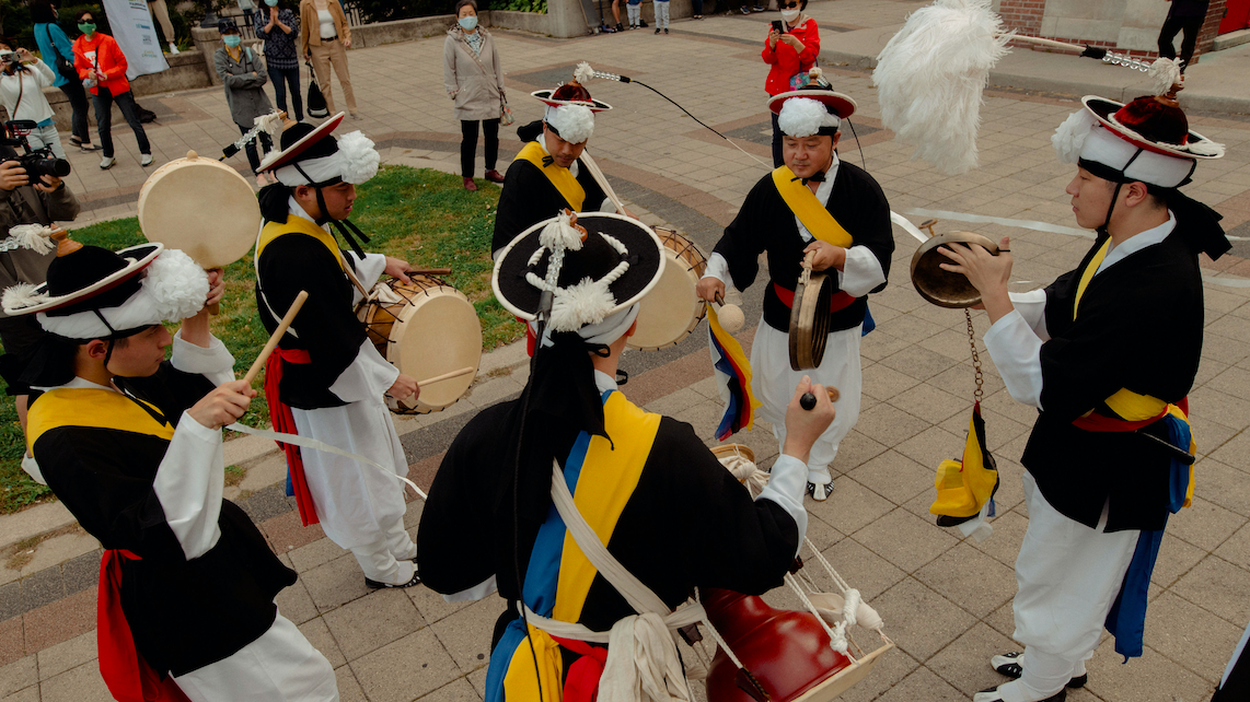 Six drummers stand in a circle in costumes. They all play handheld percussion instruments.
