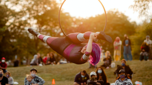 Audience members sit on the grass as a woman in costume hangs sideways from a large hoop suspended from a rope.