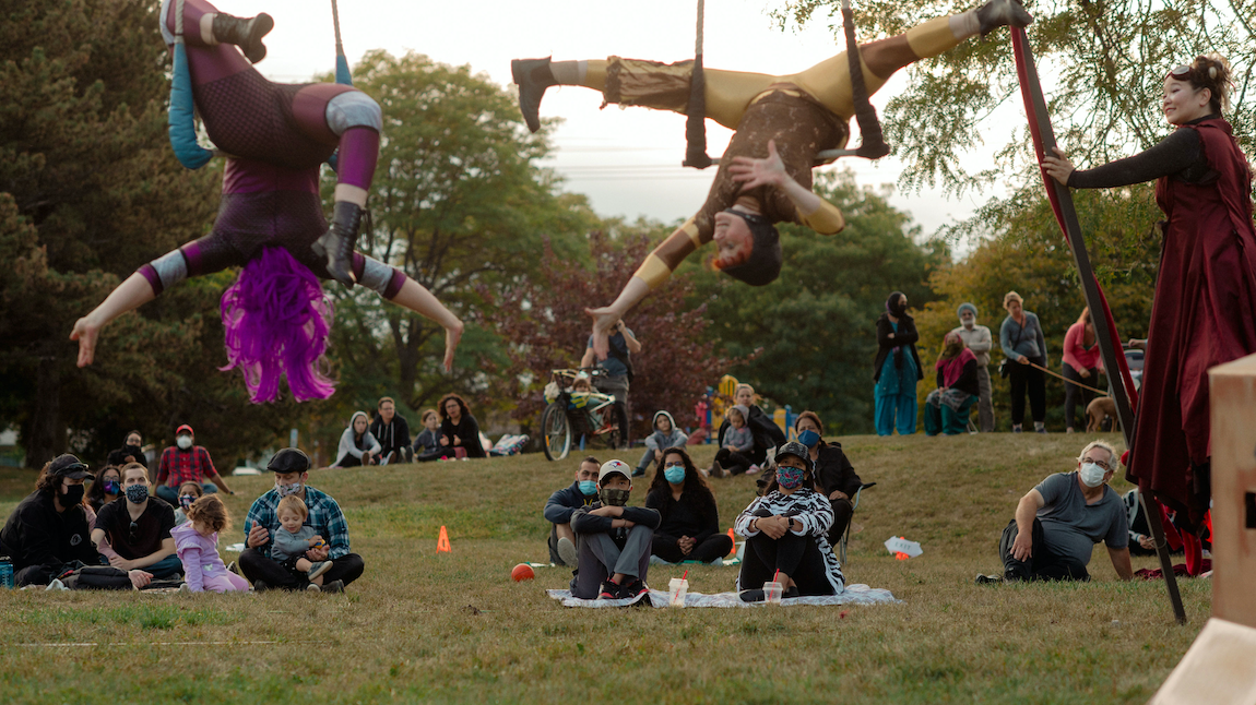 Audience members sit on the grass as two performs hang upside down in front of them.
