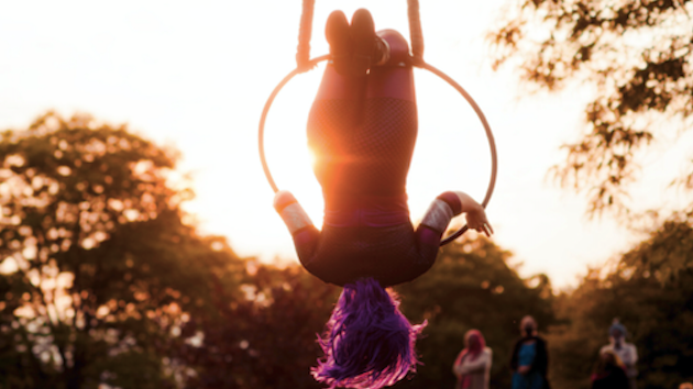 A woman in costume hangs upside down from a large hoop suspended from a rope against a background of trees and a sunset.