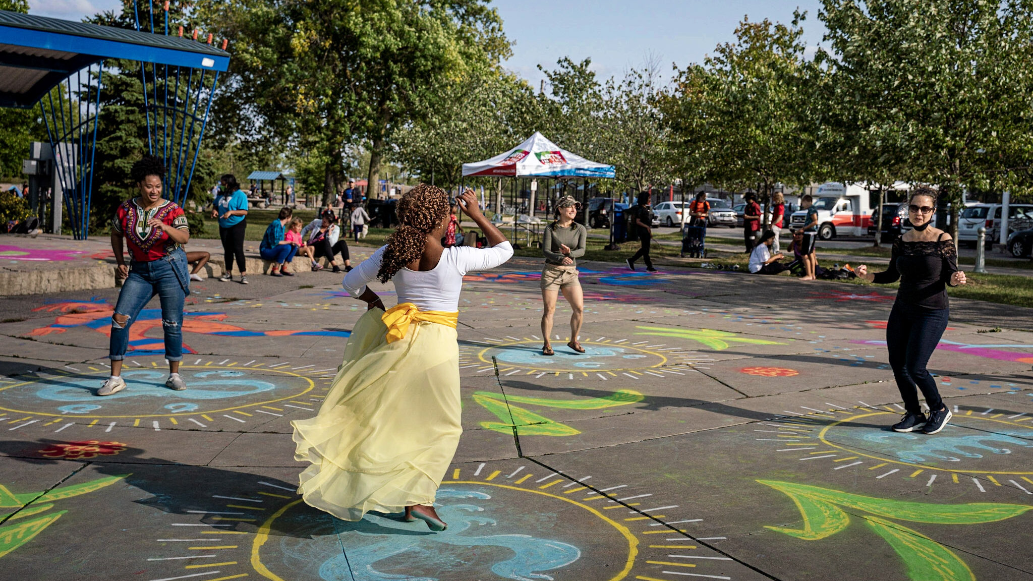 A woman in a white shirt and green skirt leads a dance workshop. Three people follow her movements.