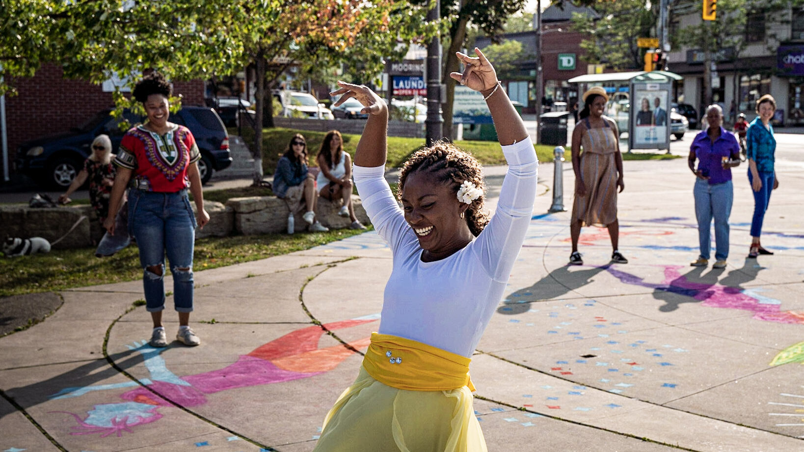 A woman in a white shirt and green skirt leads a dance workshop. Her arms are in the air and she is smiling.