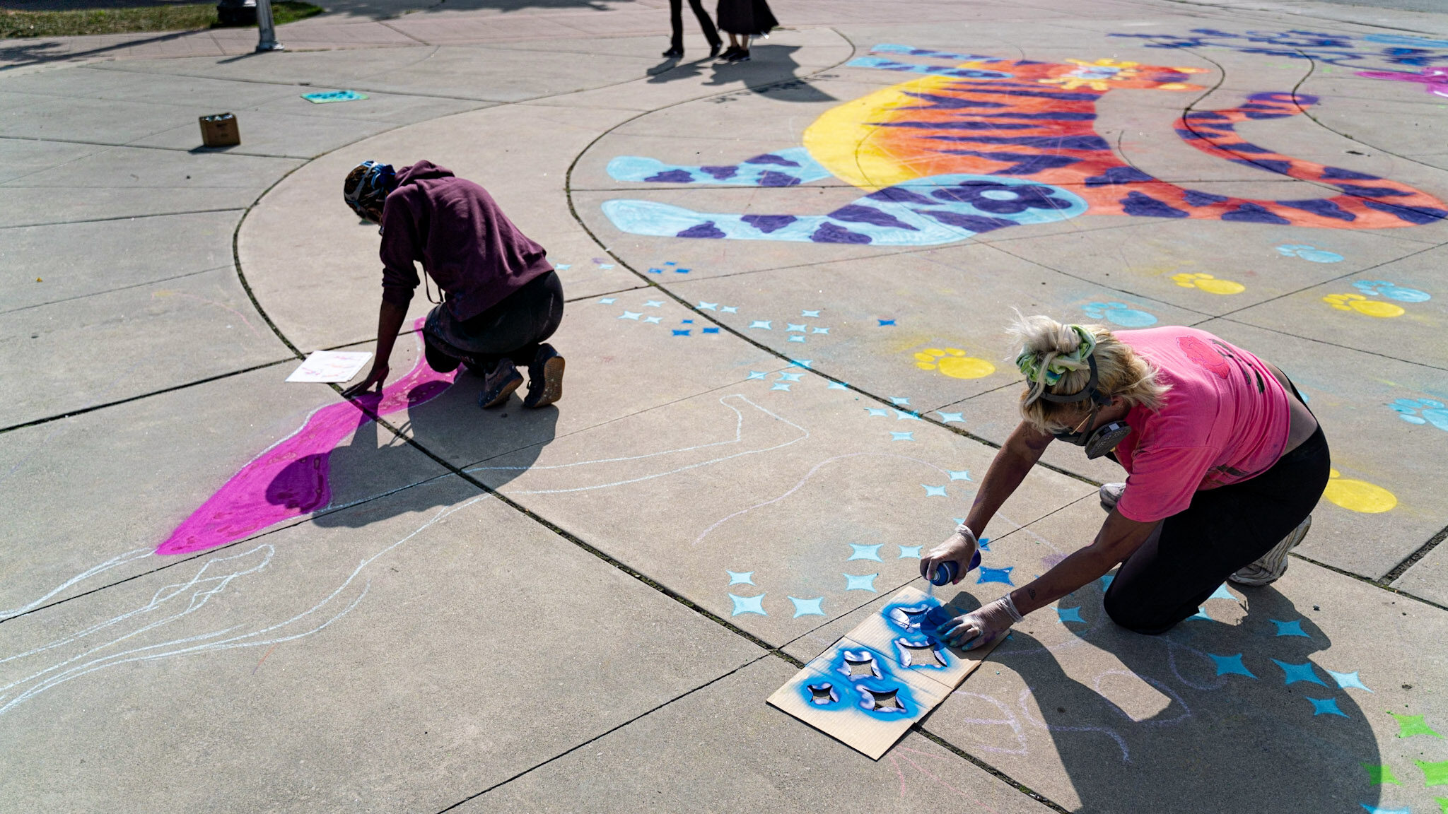 Two women are kneeling on the pavement and painting a design.
