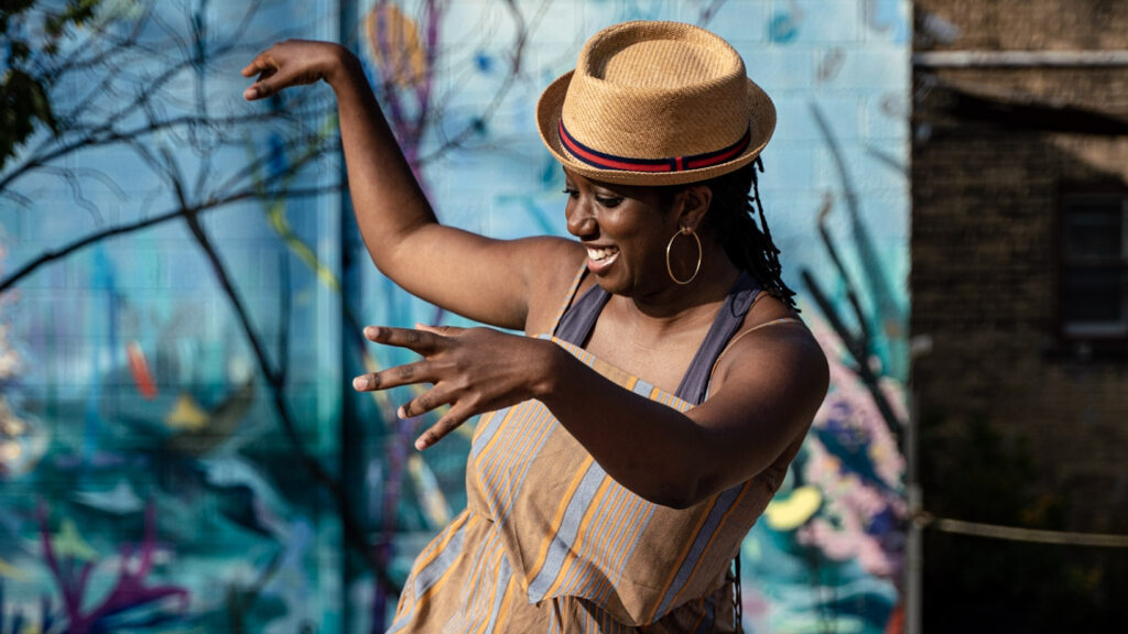 A woman dances in front of the camera, she is smiling and her arms in the air, a blue mural is in the background.