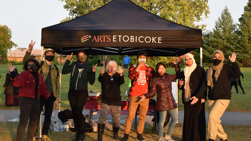 A group of adults stand in front a black tent that says "Arts Etobicoke" and wave at the camera. They are all wearing face masks.