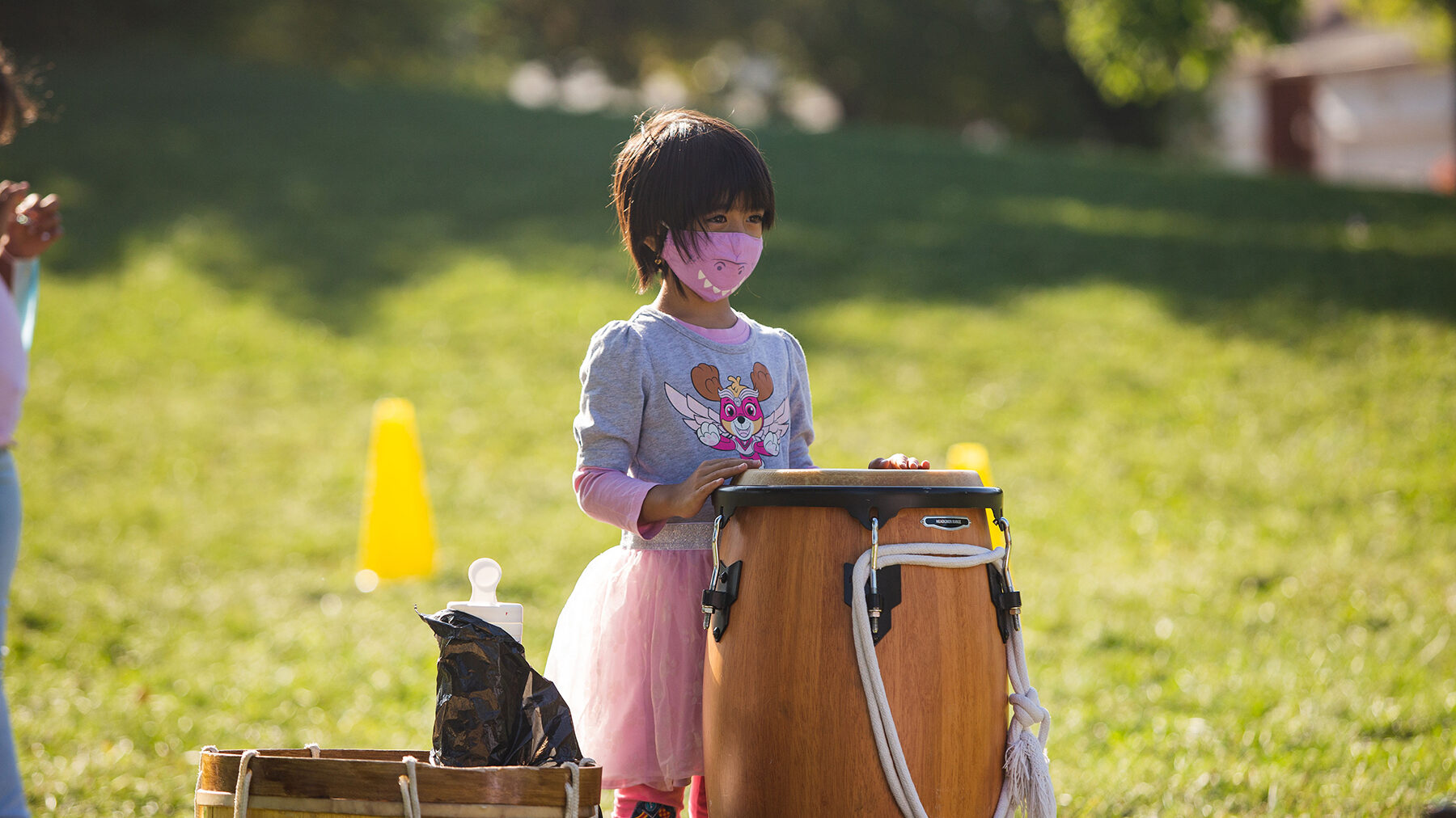 A child with a pink tutu stands in front of a large drum that is placed on the ground in front of them.