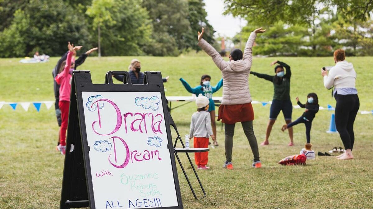 A sign in the front says "Drama Dream" while adults and children dance in a circle in the background.