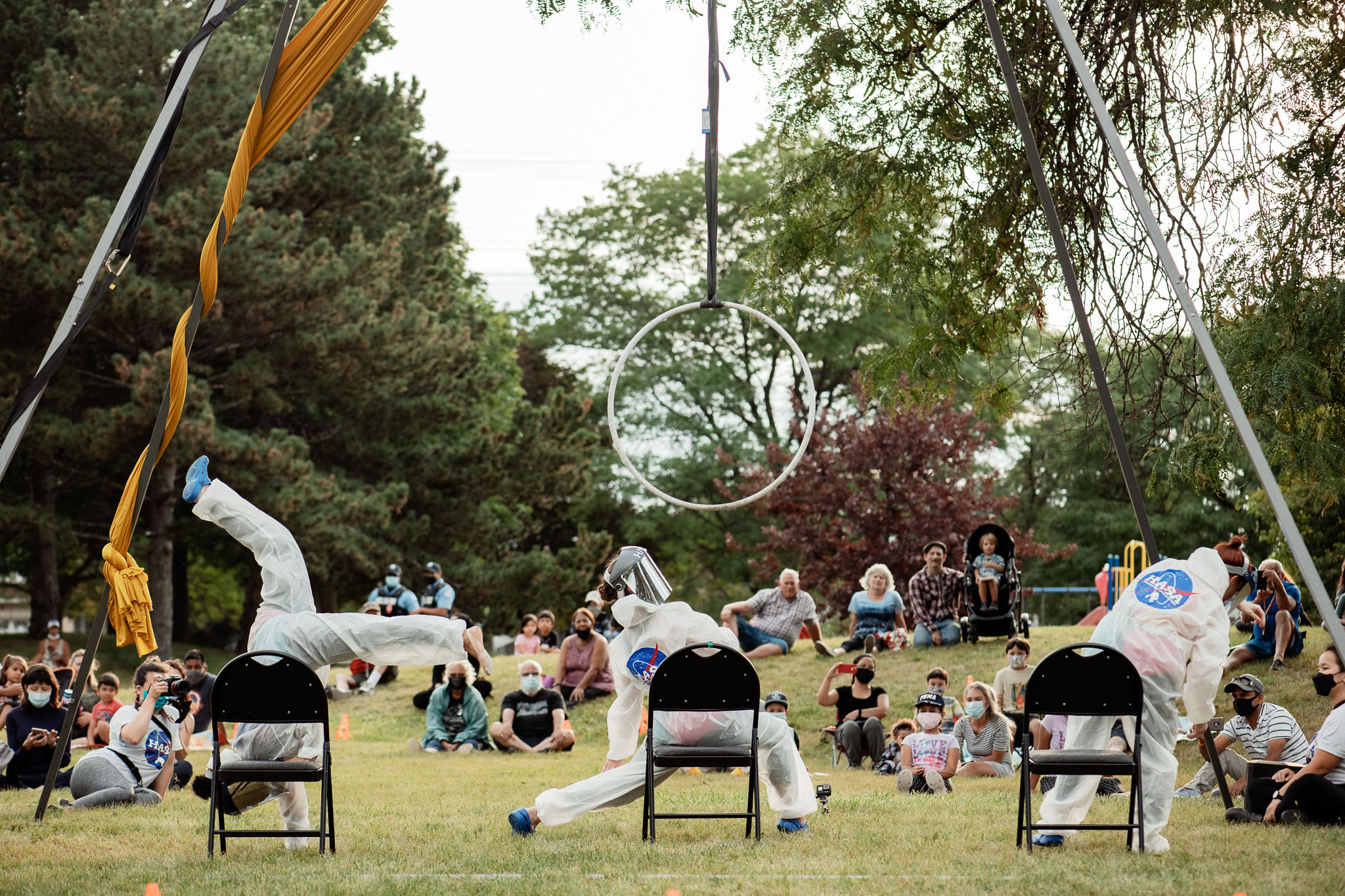 Three performers in white space suits dance on chairs while audience members watch from the grass.