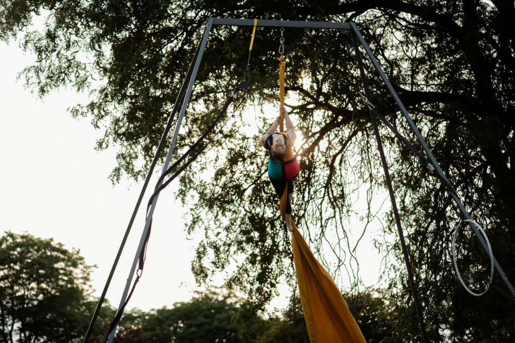A woman in a multicoloured costume hangs from an aerial rig, she is suspended by two long pieces of cloth.