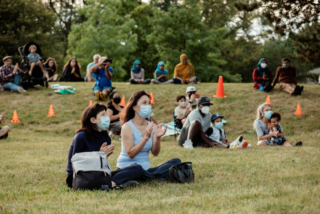 Audience members sit on the grass and clap during a performance. They are wearing face masks.