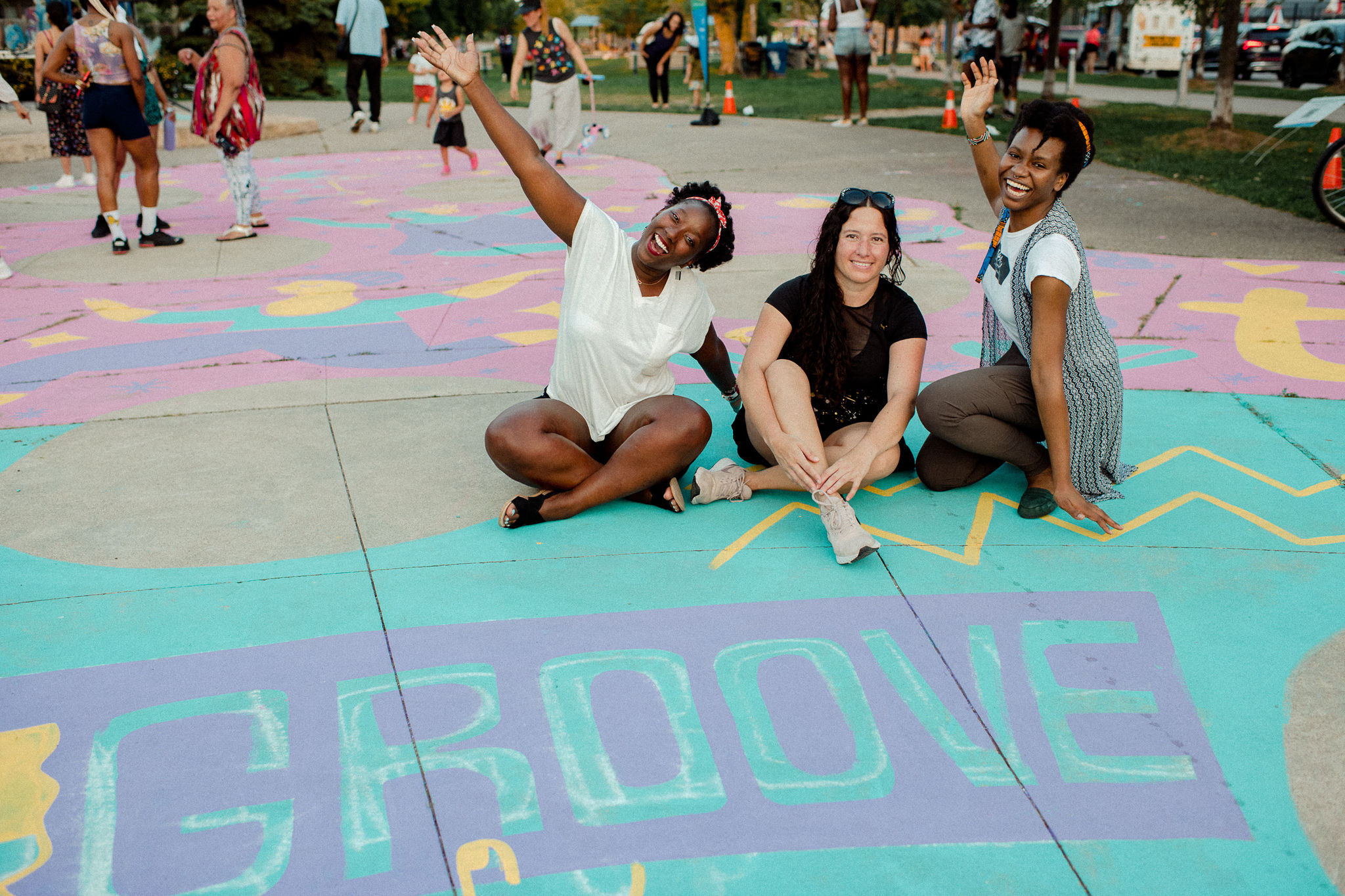 Three women sit in front of the word "groove" which is painted on pavement. They are smiling and looking directly at the camera.
