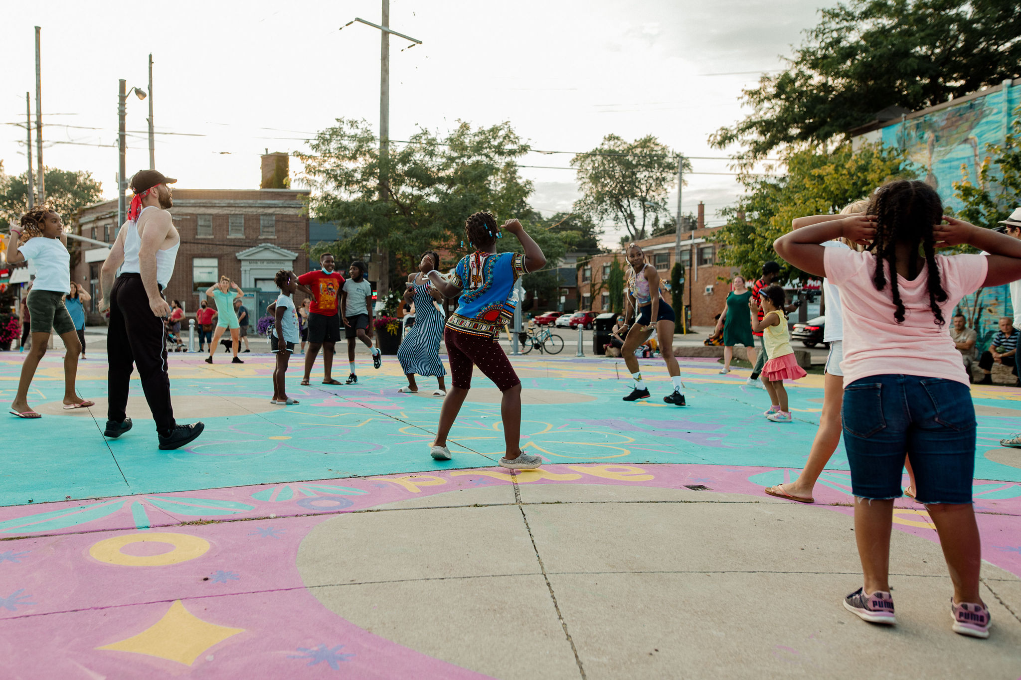 Audience members dance with their hands in the air as they participate in a workshop. They dance on brightly coloured pavement.