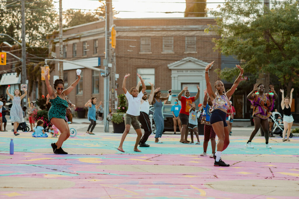 A woman dances and leads a workshop. Audience members dance behind her on brightly painted pavement.