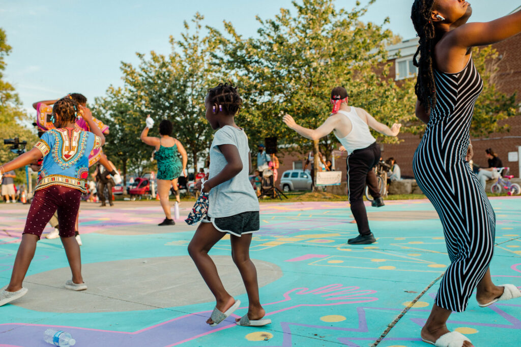 Audience members dance with their hands in the air as they participate in a workshop. They dance on brightly coloured pavement.