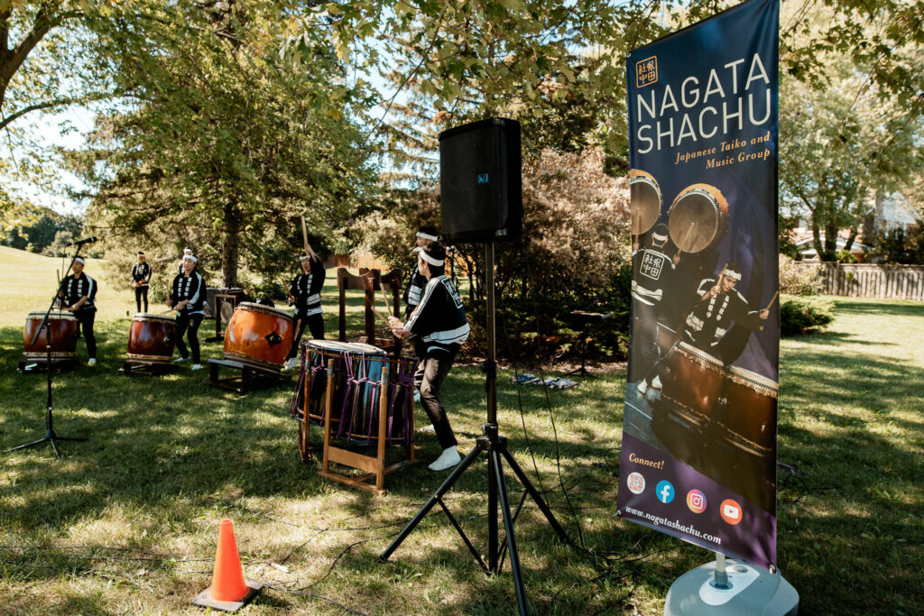 A sign with the words "Nagata Shachu" stands next to a group of drummers performing under the trees.