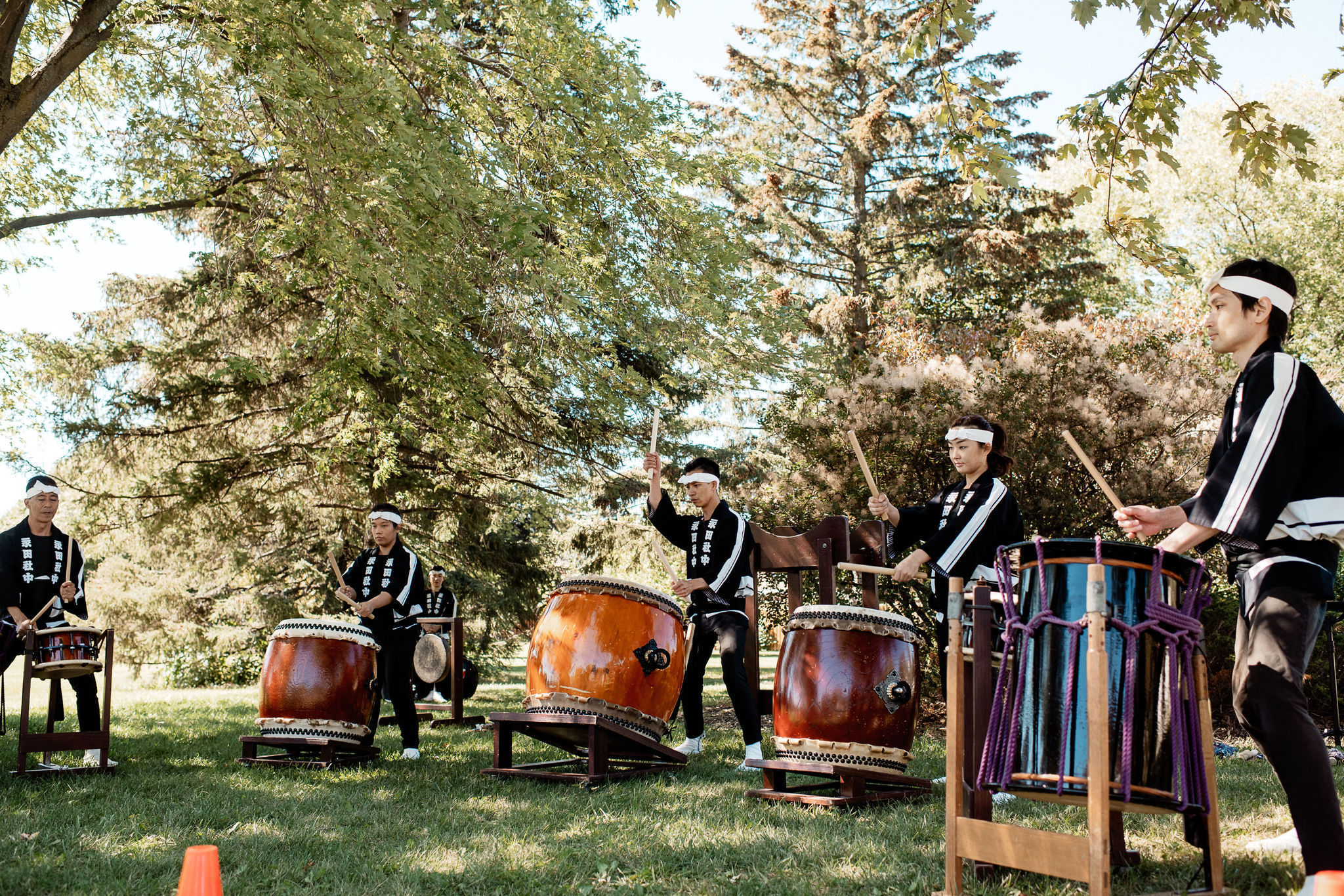 A group of five drummers play different large percussion instruments under the trees.