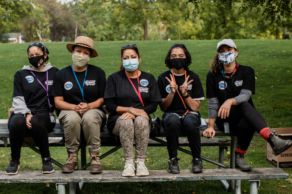 A group of people in black shirts sit on a bench and look directly at the camera. They are wearing face masks.