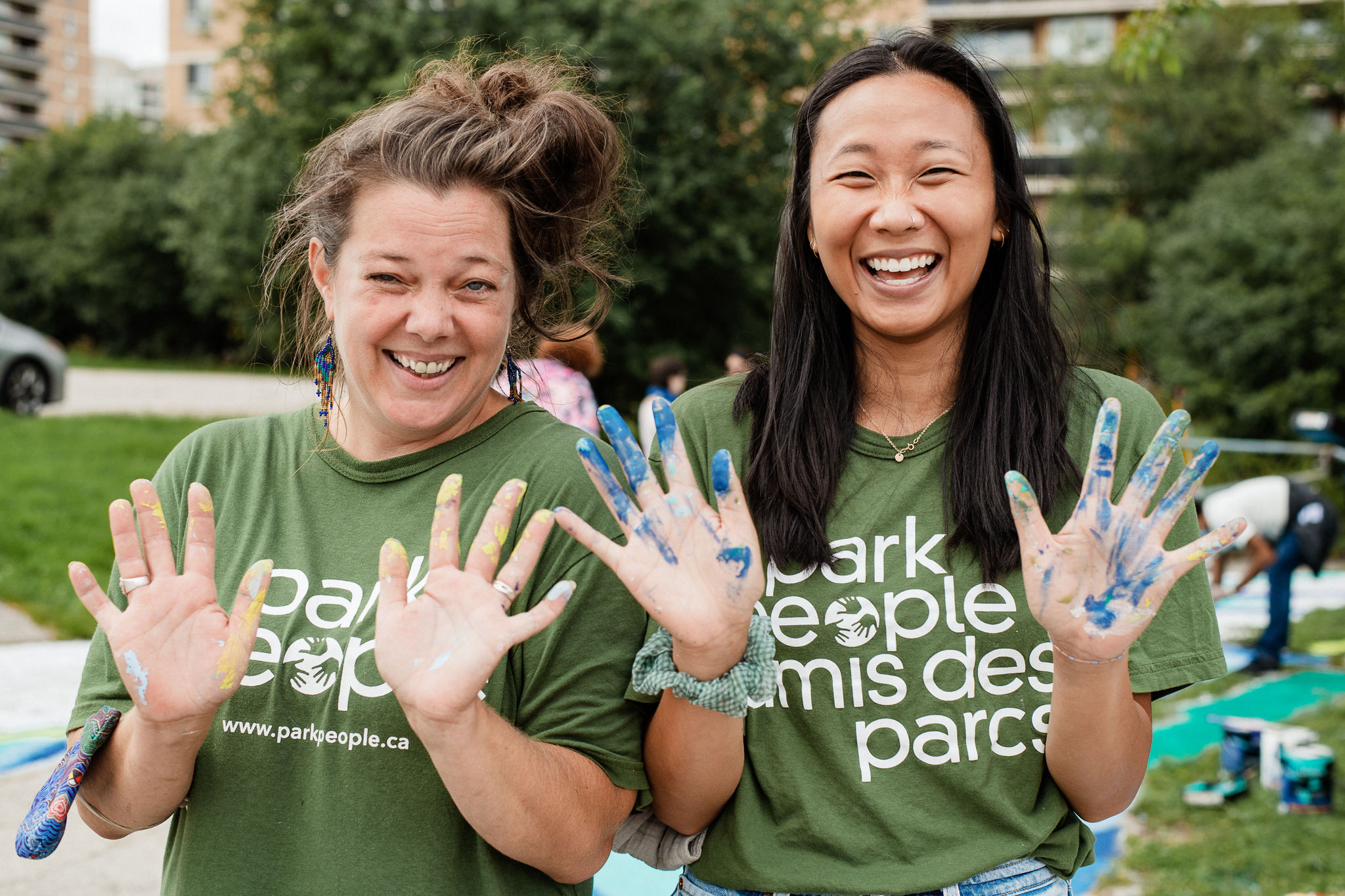 Two women wearing green shirts that say "Park People" hold up their hands and look directly at the camera while smiling.