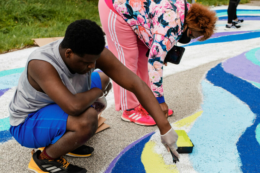 A boy and a women crouch down and paint the pavement yellow and blue.