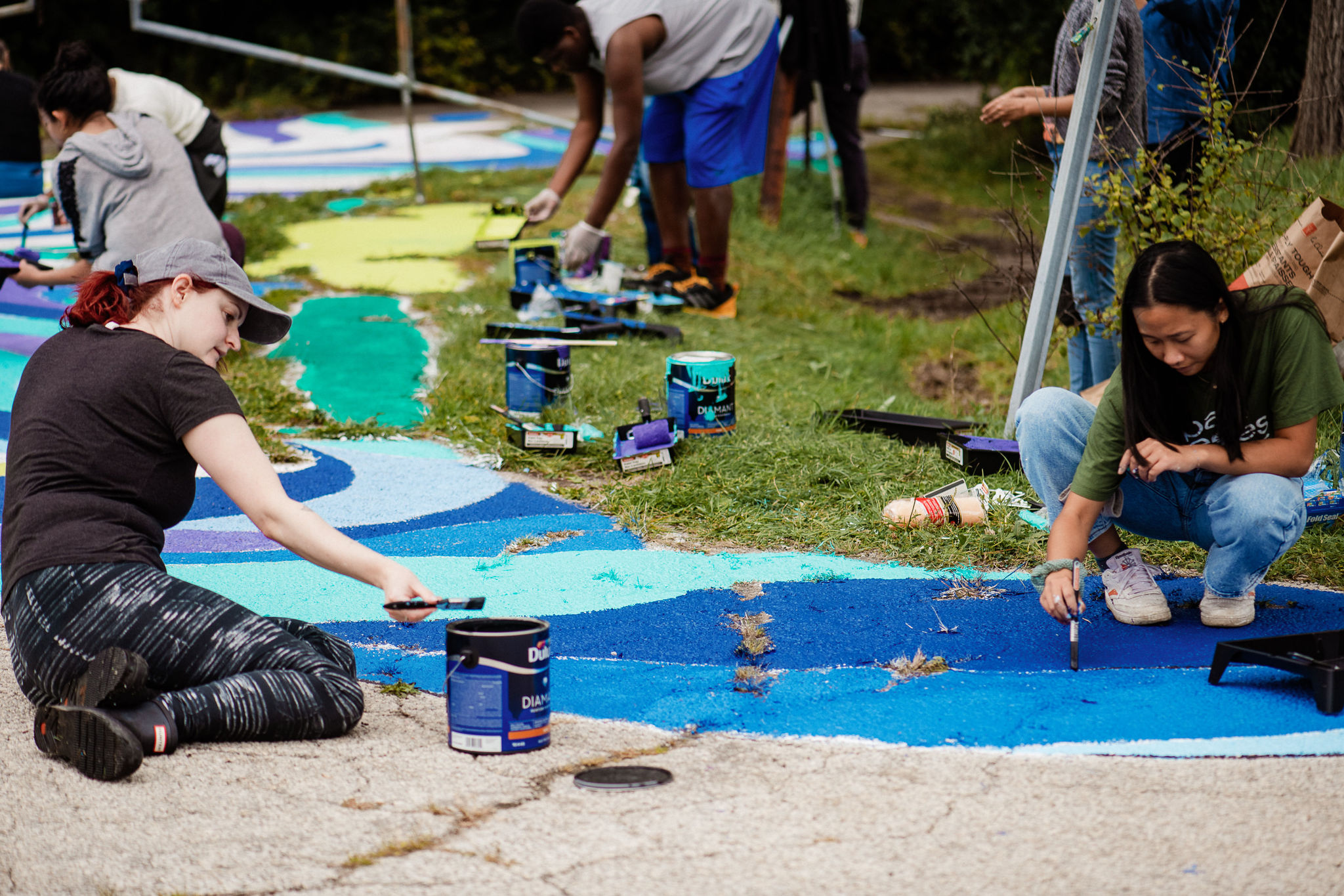 A group of four people sit on the ground and paint the pavement different shades of blue.
