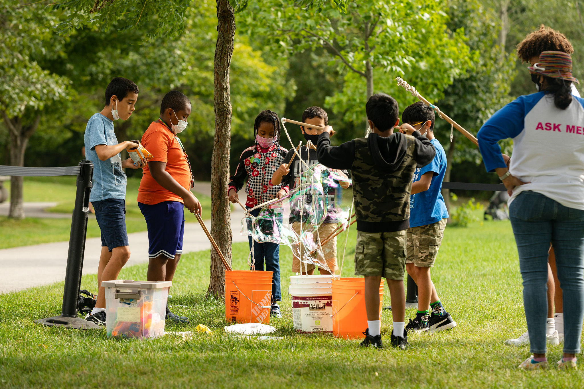 A group of children stand around a bucket and use sticks and ropes to make large bubbles.