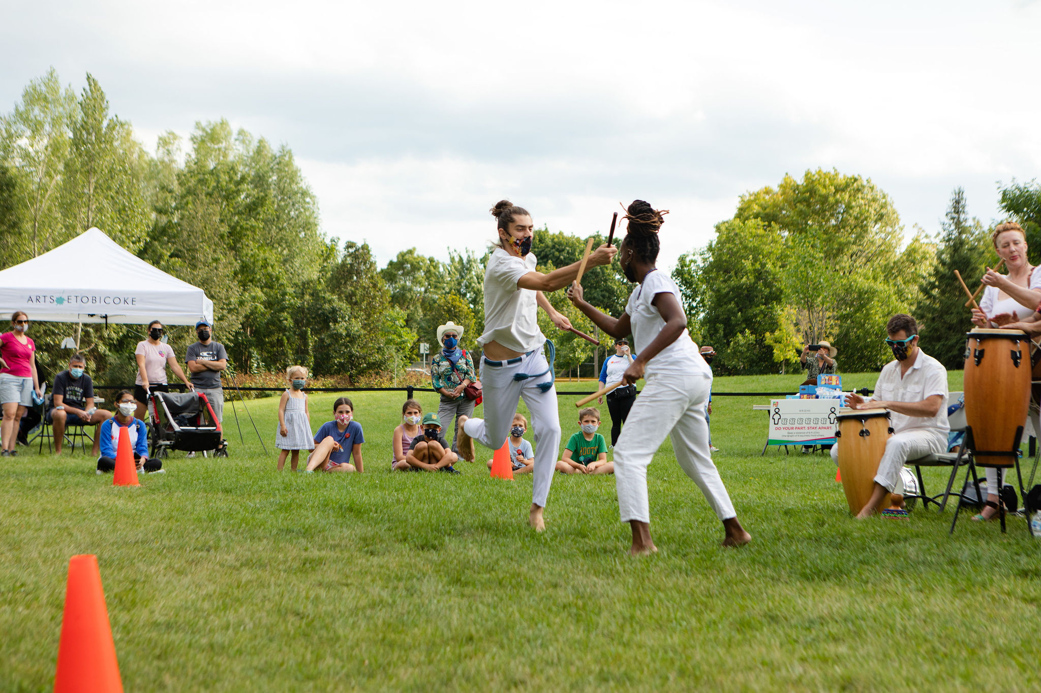 A man and woman, both in white dance together in a field while audience members watch.