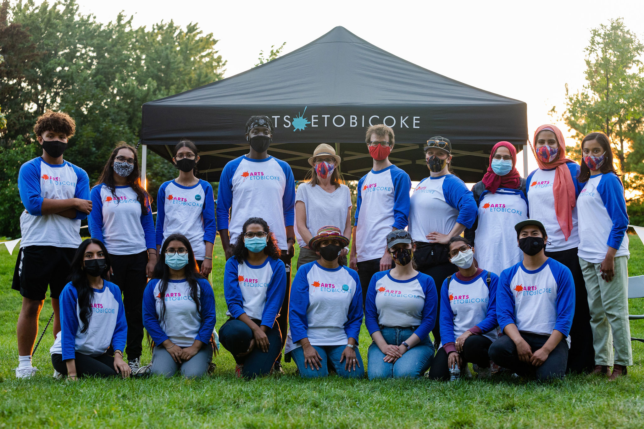 A group of people in white and blue shirts crouch and stand together in front of an Arts Etobicoke tent. They look directly at the camera and are wearing face masks.