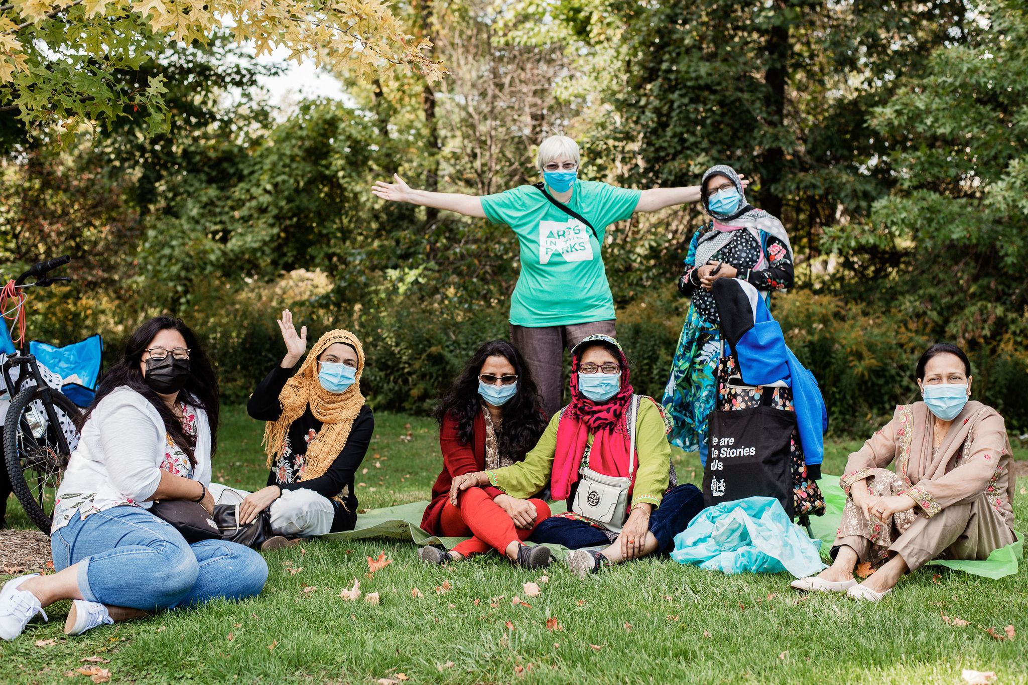 A group of women stand and sit together on the grass. They look directly at the camera, they are wearing face masks.