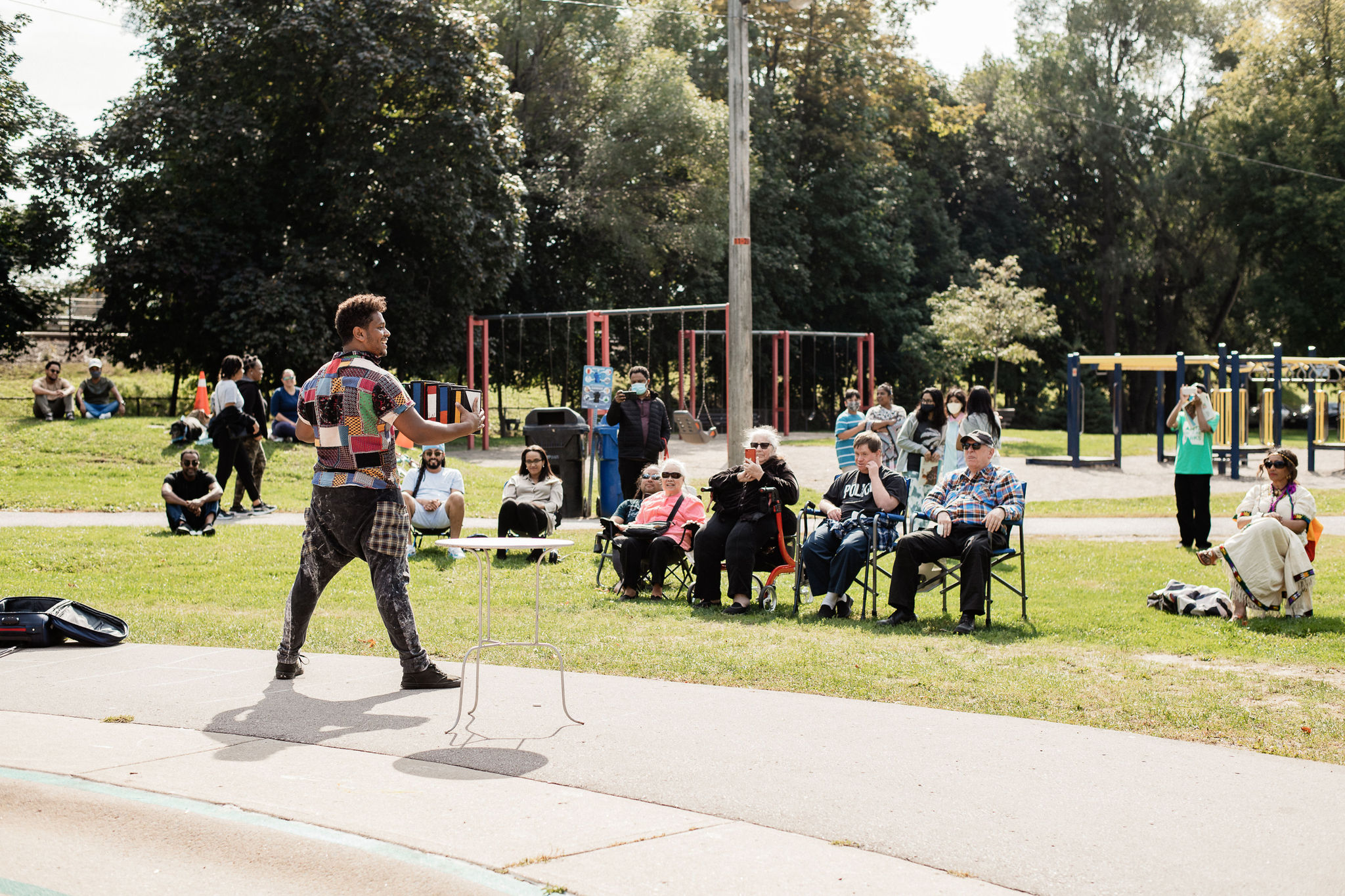 A man stands on the pavement and juggles while audience members watch from the grass.