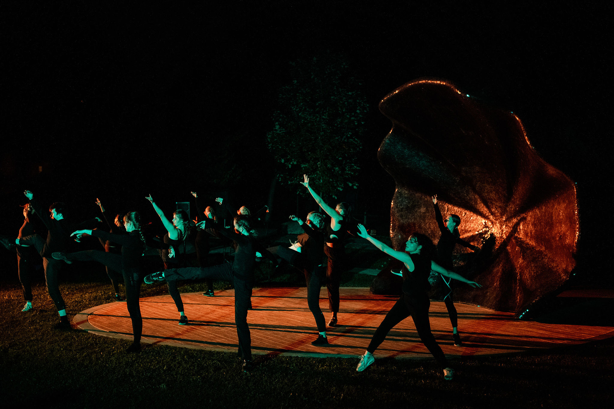 A group of dances all wearing black dance in unison on a wooden stage. They are lit with green light.